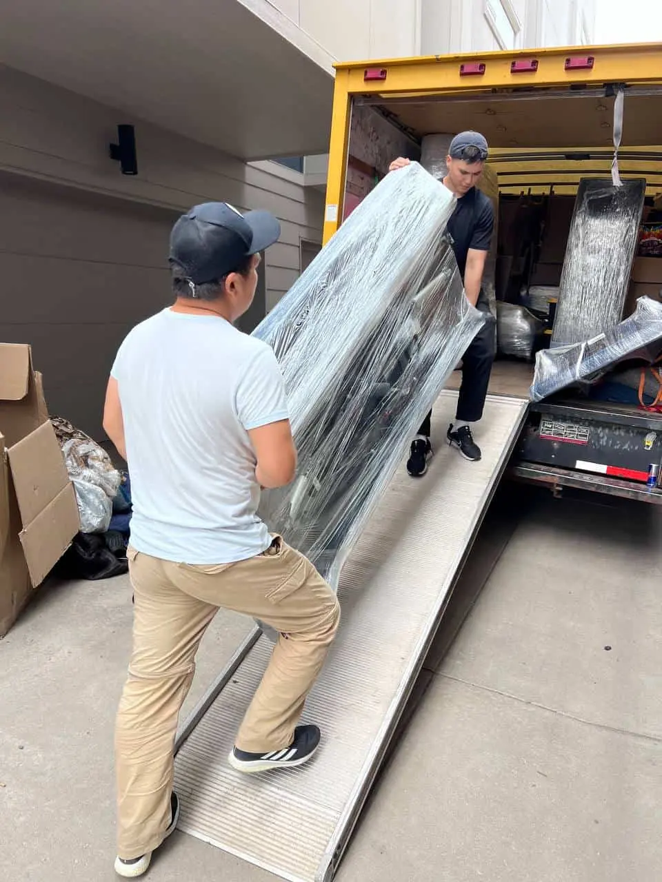 Two movers carrying a wrapped mattress up the truck ramp